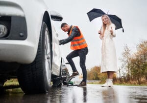 Beautiful young woman with umbrella giving thumbs up and smiling while roadside assistance worker inflating tire with air compressor. Auto mechanic repairing car while woman showing approval gesture.