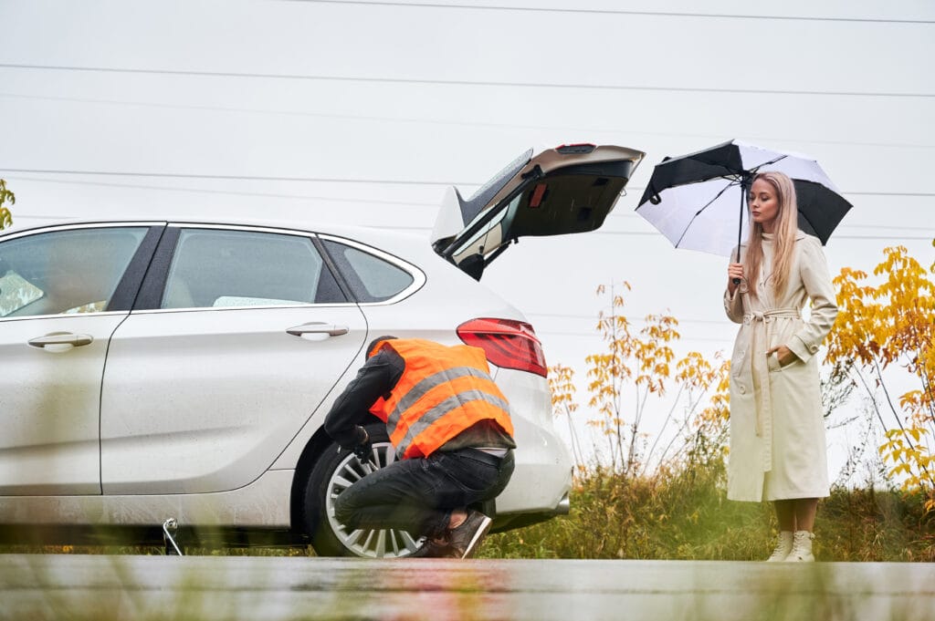 Beautiful woman holding umbrella and looking at roadside assistance worker while man unscrewing lug nuts on car wheel. Auto mechanic repairing woman car on the road. Concept of emergency road service.